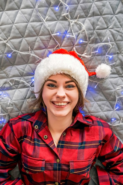 サンタの帽子をかぶった笑顔の女の子と花輪の灰色の背景に横たわっているカメラを見ている赤いパジャマの格子縞のシャツの上からのクリスマスビュー
