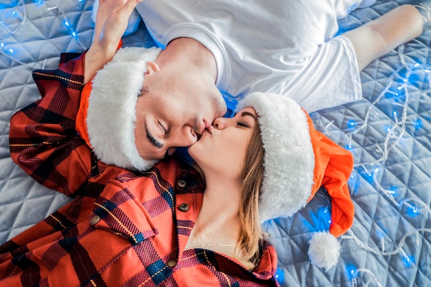 Christmas view from above on a young kissing heterosexual couple wearing santa hats and pajamas lying on bed with garland