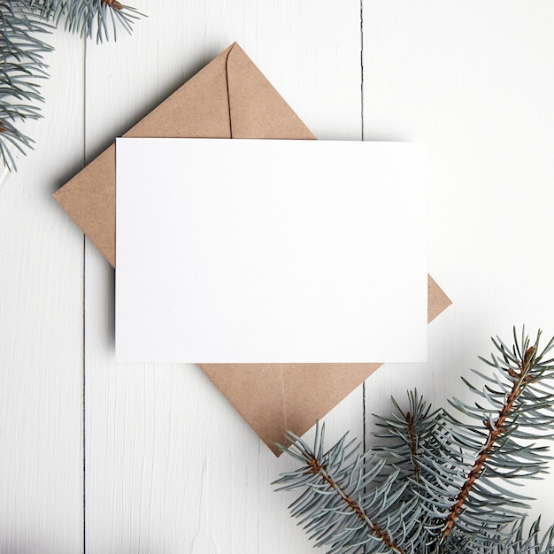 Christmas wenskaart mockup met ambachtelijke papieren envelop en verse groene dennenboomtak op witte houten achtergrond bovenaanzicht plat lag Lege wintervakantiekaart