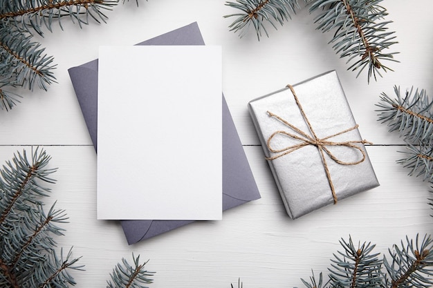 Christmas wenskaart mockup met grijze envelop zilveren kleur geschenkdoos en groene dennenboomtak op witte houten achtergrond bovenaanzicht plat lag Lege wintervakantiekaart