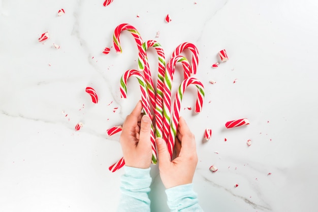 Christmas, winter concept. Holidays, sweets, treats. Girl hands holding traditional candy cane in the form of a bouquet, whole and broken into pieces. White marble table, top view,  copyspace