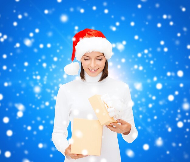 christmas, winter, happiness, holidays and people concept - smiling woman in santa helper hat opening gift box over blue snowy background