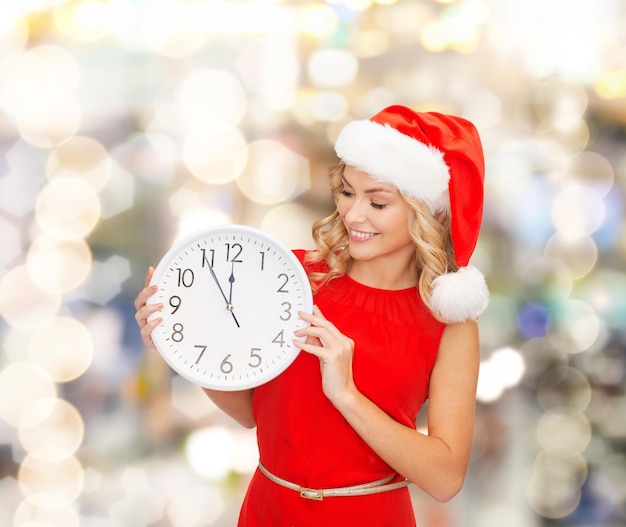 рождество, зима, праздники, время и концепция людей - улыбающаяся женщина в шляпе помощника Санты и красном платье с часами на фоне огней