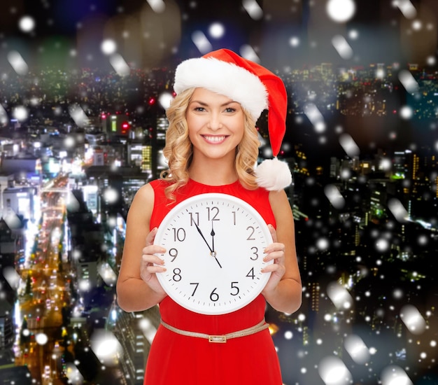 christmas, winter, holidays, time and people concept - smiling woman in santa helper hat and red dress with clock over snowy city background