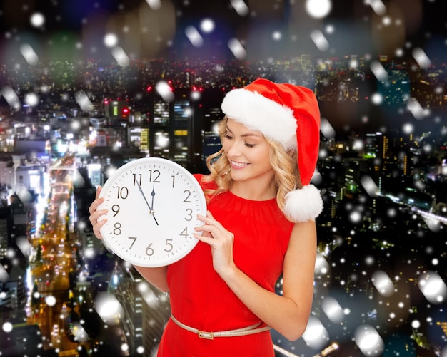 クリスマス、冬、休日、時間と人々の概念-雪の降る夜の街の背景に時計とサンタヘルパー帽子と赤いドレスの笑顔の女性