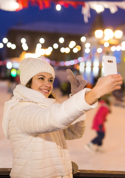 Christmas, winter, technology and leisure concept - happy young woman taking picture with smartphone on ice skating rink outdoors.