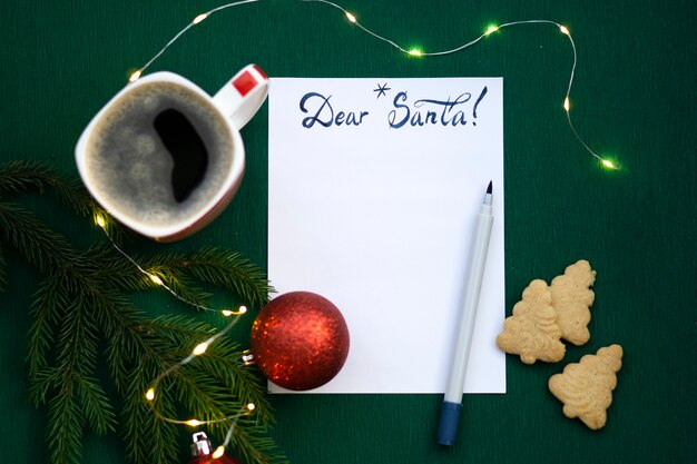 Christmas wishlist 2022 or letter to Santa on green colored paper background. Festive decoration, Christmas garland, Christmas tree branch. Flat lay. View from above.