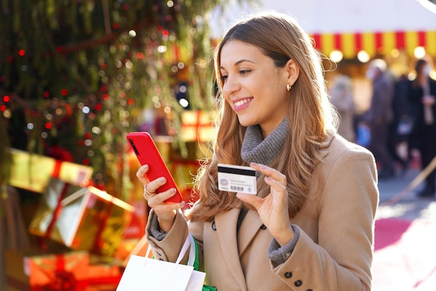 Christmas woman carrying shopping bags outdoor beautiful\
smiling girl paying her gifts online with mobile phone and credit\
card with blurred people on christmas markets in the\
background