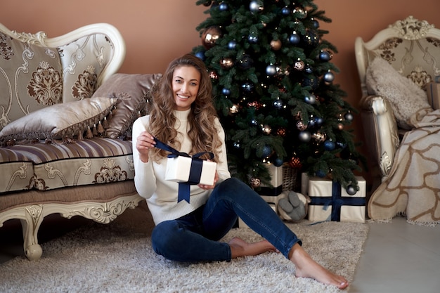 Christmas. Woman dressed white sweater and jeans sitting on the floor near christmas tree with present box