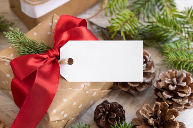 Foto regalo di natale avvolto con etichetta regalo di carta con rami di abete, pigne e decorazioni natalizie da vicino. composizione invernale rustica con etichetta regalo vuota mockup, copia spazio, primo piano