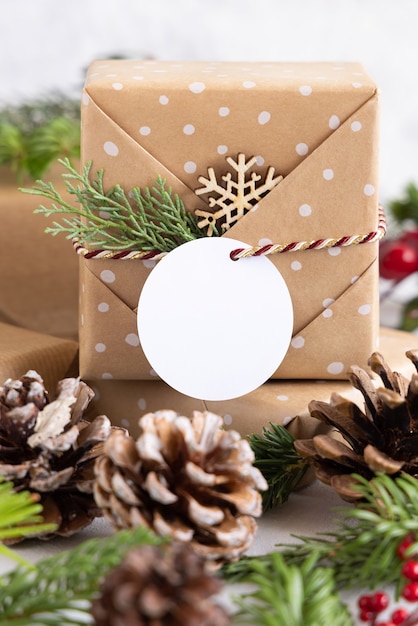Christmas wrapped present with round paper gift tag on a white table with fir tree branches and decorations close up. Rustic winter composition with blank Gift tag Mockup, copy space