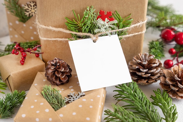 Christmas wrapped present with square paper gift tag on a white table with fir tree branches and decorations close up. Rustic winter composition with blank Gift tag Mockup, copy space