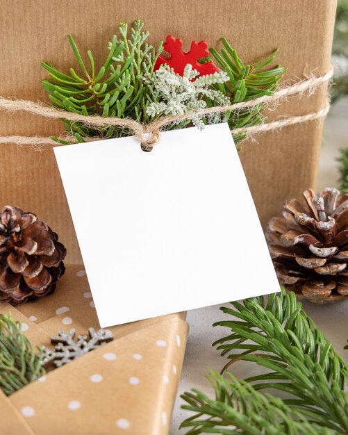 Christmas wrapped present with square paper gift tag on a white table with fir tree branches and decorations close up. Rustic winter composition with blank Gift tag Mockup, copy space