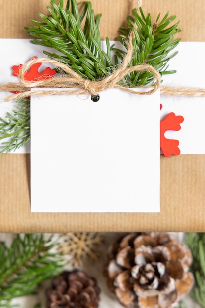 Christmas wrapped present with square paper gift tag on a white table with fir tree branches and decorations top view. Rustic winter composition with blank Gift tag Mockup, copy space