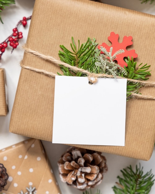 Christmas wrapped present with square paper gift tag on a white table with fir tree branches and decorations top view. Rustic winter composition with blank Gift tag Mockup, copy space