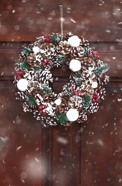 Christmas wreath decorated with bumps and berries hanging on a wooden door during the snowfall. Closeup shot