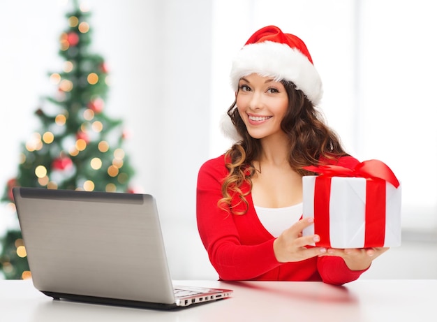 christmas, x-mas, online shopping concept - woman in santa helper hat with gift box and laptop computer