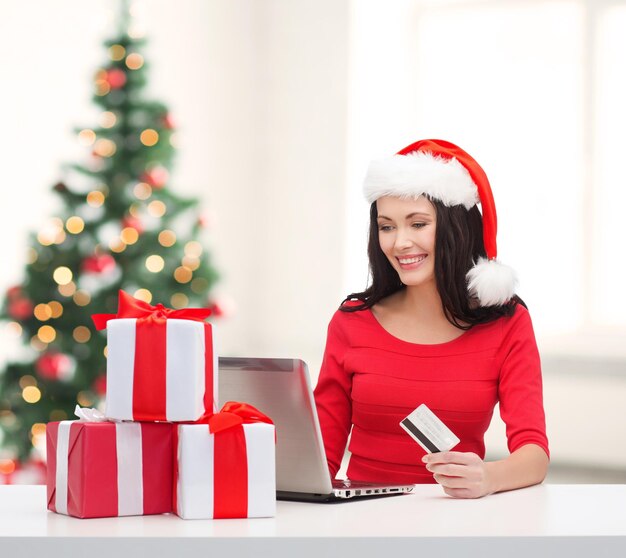 christmas, x-mas, online shopping, electronics concept - woman in santa helper hat with gift boxes, laptop computer and credit card