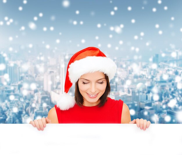 크리스마스, 크리스마스, 사람, 광고 및 판매 개념 - 눈 덮인 도시 배경 위에 빈 흰색 보드가 있는 산타 도우미 모자를 쓴 행복한 여성