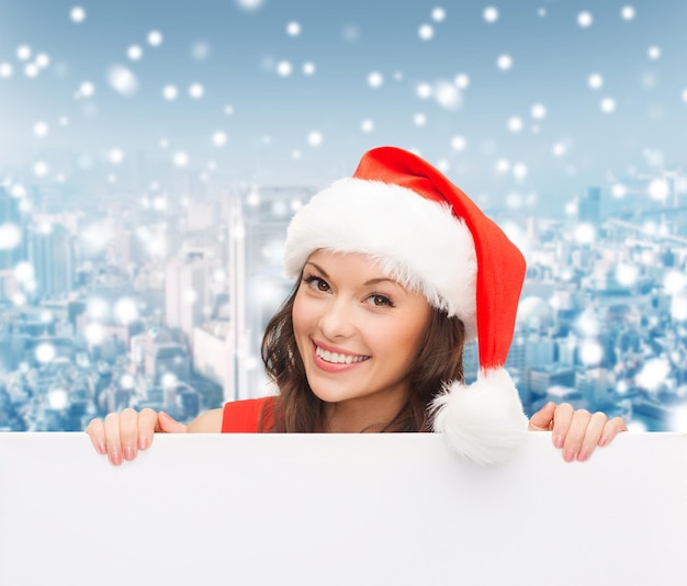 christmas, x-mas, people, advertisement and sale concept - happy woman in santa helper hat with blank white board over snowy city background