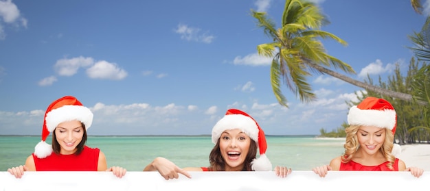 Photo christmas, x-mas, people, advertisement and sale concept - happy women in santa helper hat with blank white board over tropical beach background