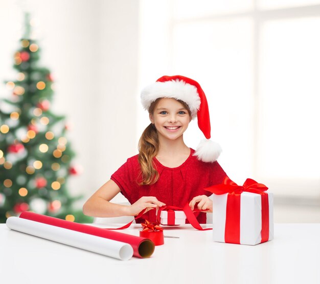 christmas, x-mas, winter, happiness concept - smiling girl in santa helper hat with gift box and wrapping paper