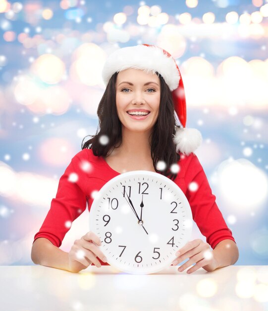 Christmas, x-mas, winter, happiness concept - smiling woman in santa helper hat with clock showing 12