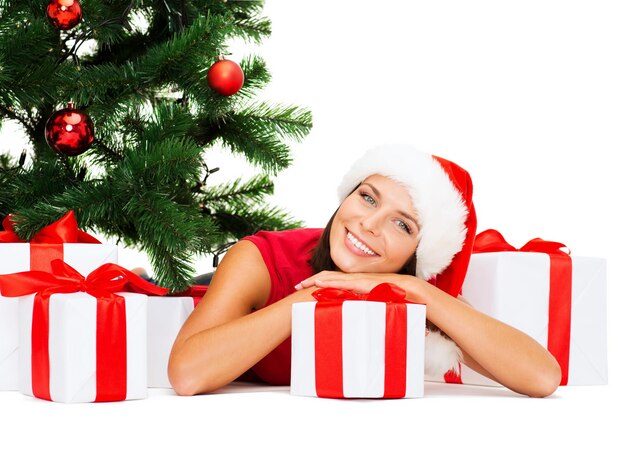 christmas, x-mas, winter, happiness concept - smiling woman in santa helper hat with many gift boxes and tree