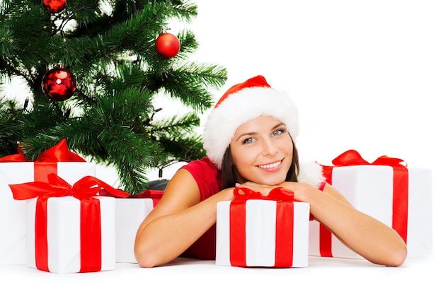 christmas, x-mas, winter, happiness concept - smiling woman in santa helper hat with many gift boxes and tree