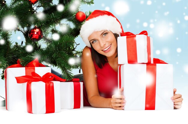 christmas, x-mas, winter, happiness concept - smiling woman in santa helper hat with many gift boxes and tree