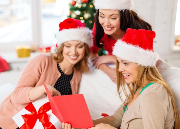 christmas, x-mas, winter, happiness concept - three smiling women in santa helper hats with card and many gift boxes