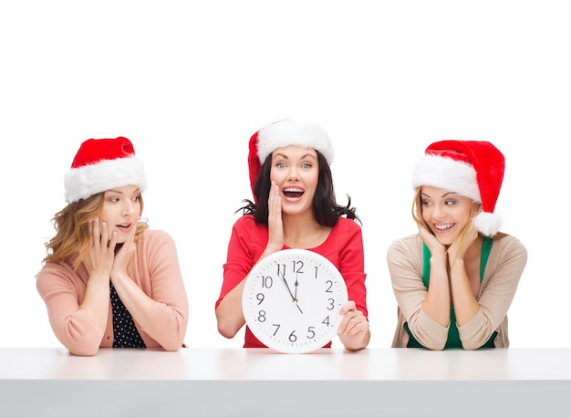 рождество, рождество, зима, концепция счастья - три улыбающиеся женщины в шляпах санта-помощника с часами, показывающими 12