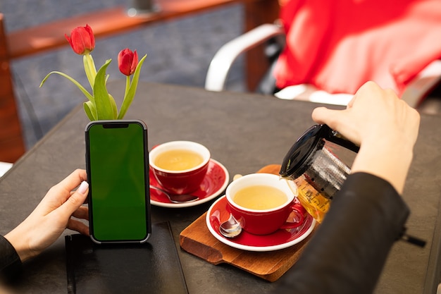 Chromakey telefoonscherm in handen van vrouw die thee drinkt in café met kopieerruimte, lifestyle