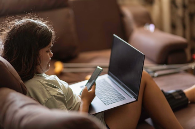 Photo chubby asian teen girs with injured leg and crutches talking on the phone and laptop at home