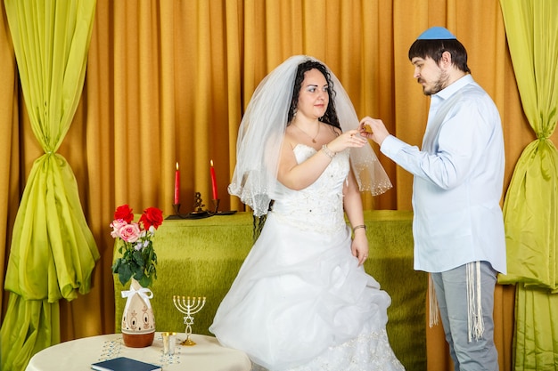 During a chuppah ceremony at a Jewish synagogue wedding, the groom puts a ring on the bride's index finger. Horizontal photo