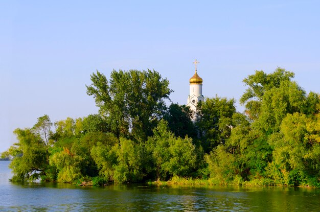 Church in the trees on the bank of the river, blue sky and reflection in water