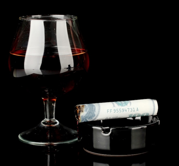 Сигара 100-долларовой банкноты и пепельница и стакан бренди, изолированные на черном