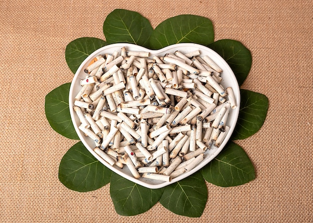 Cigarette butts on a white plate in the shape of a heart. The plate lies on the leaves. Burlap background.