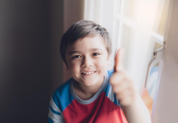 Кинематографический портрет Счастливый мальчик смотрит в камеру с улыбающимся лицом Позитивный ребенок сидит рядом с окном в гостиной и отдыхает на выходных весной или летом