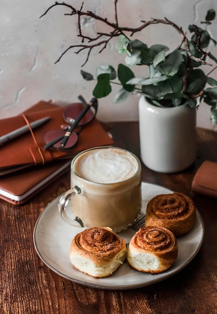 Cinnamon puff pastry buns cappuccino vase with decorative flowers books on a wooden table in a cozy room