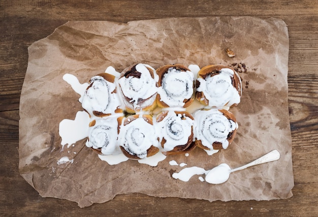 Cinnamon rolls with cream cheese icing on piece of oily craft paper over rustic wooden 