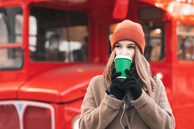 Citeer dame in een warme jas en oranje hoed, staande op straat op de achtergrond van een rode bus, koffie drinkend uit een papieren bekertje, zijwaarts kijkend