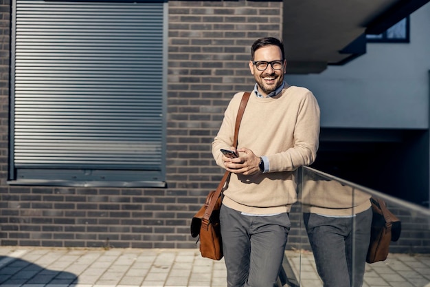 A city man leaning on the glass outside typing messages on the phone and smiling at the camera