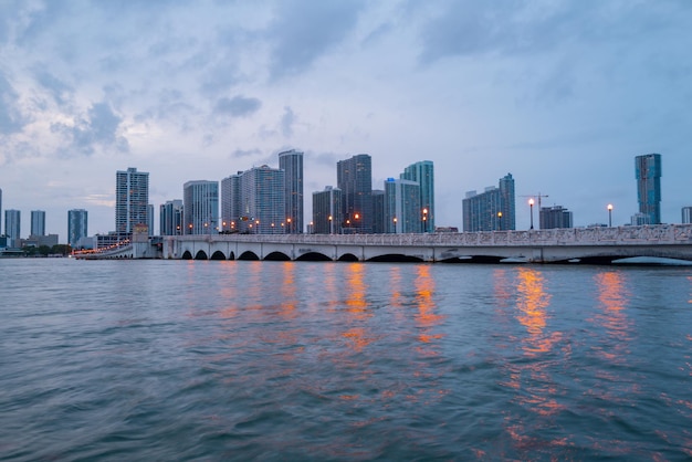 Город Майами Флорида, панорама заката с деловыми и жилыми зданиями и мостом на заливе Бискейн. Ночной вид на горизонт.
