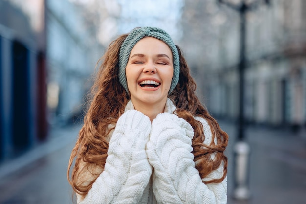 A city portrait on a blurry background with a young great female in a white knitted look for cold weather. Showing a insane happiness. A girl laughing loudly. The concept of happiness.