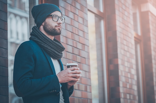 シティスタイル。コーヒーカップを保持し、通りに立っている間目をそらしているスマートカジュアルな服装のハンサムな若い男