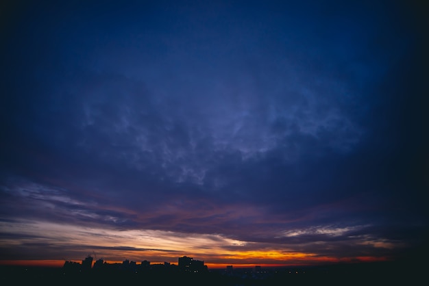 Cityscape with vivid warm dawn. Amazing dramatic blue violet cloudy sky above dark silhouettes of city buildings. Orange sunlight. Atmospheric background of sunrise in overcast weather. Copy space.