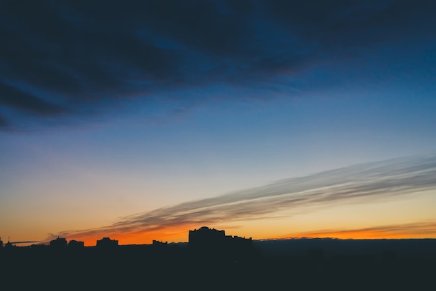 Cityscape with wonderful varicolored vivid dawn. Amazing dramatic blue cloud sky above dark silhouettes of city building roofs