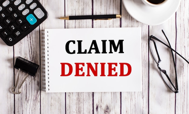 CLAIM DENIED is written in a white notepad near a calculator, coffee, glasses and a pen. Business concept