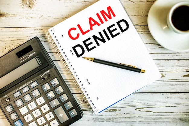 CLAIM DENIED written on white paper near coffee and calculator on a light wooden table